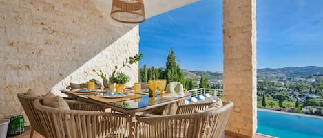 A dining area with wicker furniture, a table set with plates, glasses, and a hat, and a view of a pool and landscape.