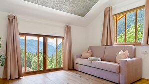 A room with a sofa, a large window with curtains, wooden flooring, and a view of mountains.