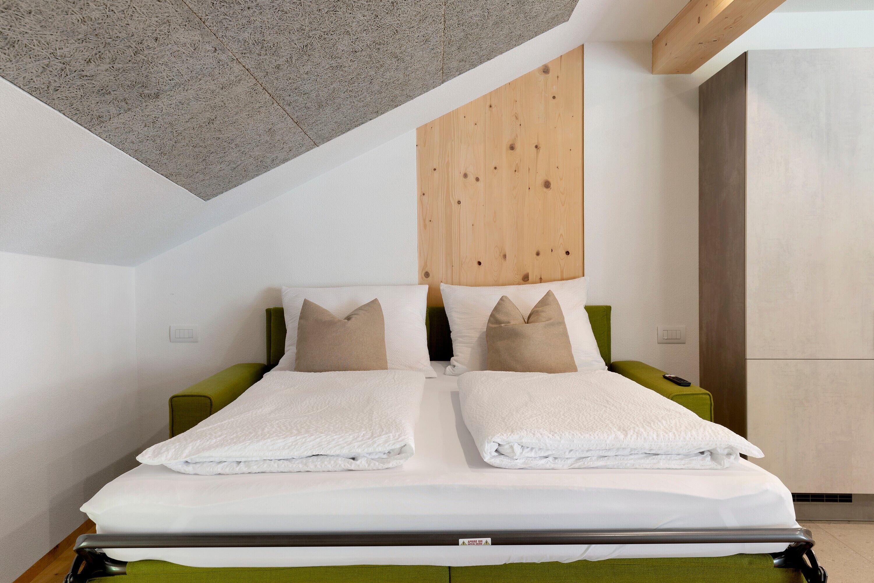 A modern hotel room with a sloped ceiling, a wooden headboard, a bed with white linens, and a green upholstered bench.