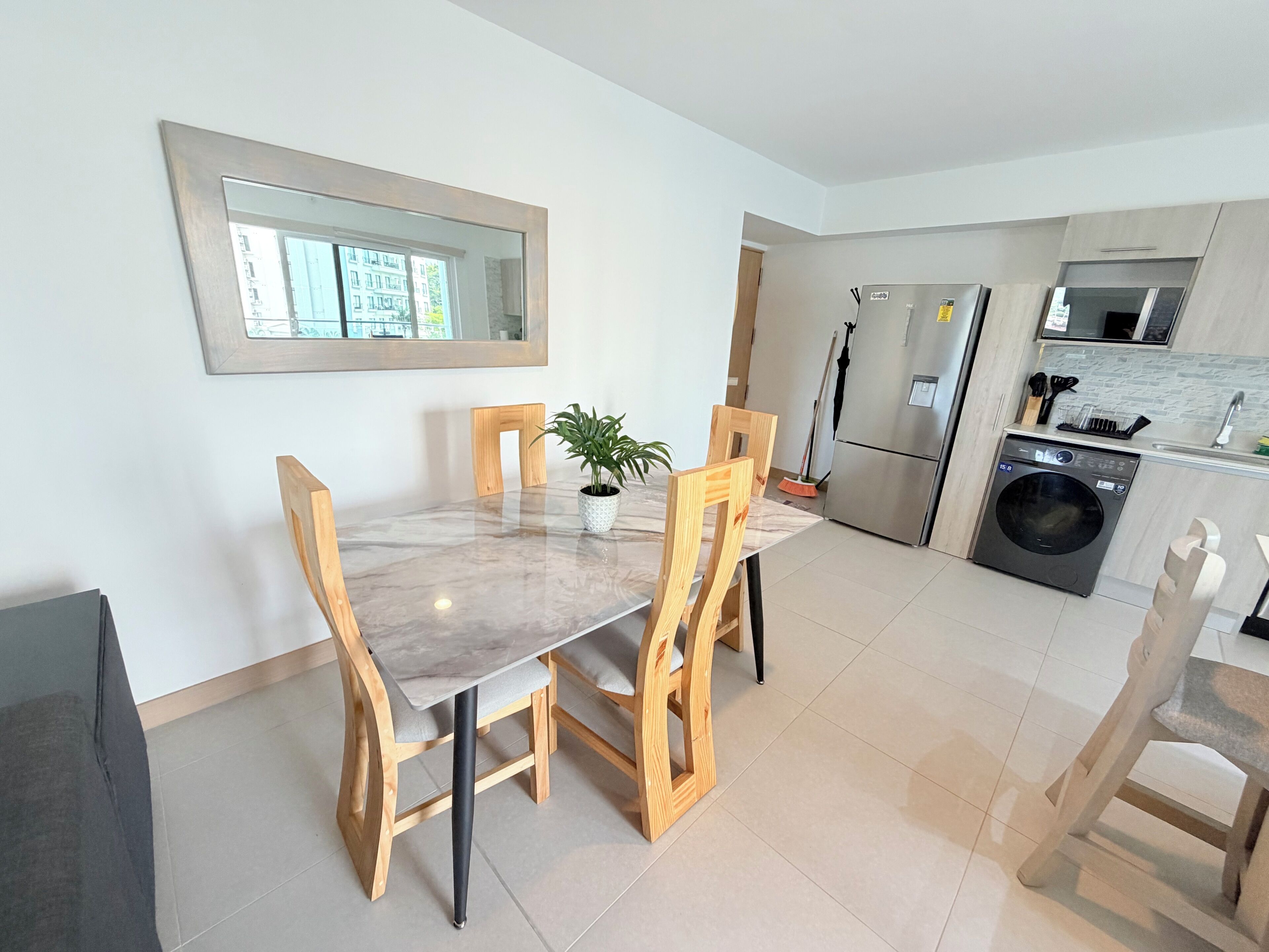 A modern kitchen with a dining table, chairs, a refrigerator, a washing machine, and a window with a view of buildings.