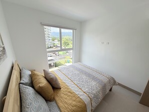 Ein Schlafzimmer mit Bett, Kissen, einem Fenster und Blick auf Gebäude und Grünflächen.