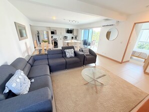 Ein modernes Wohnzimmer mit einem Ecksofa, einem Glastisch und Blick auf die Küche sowie den Außenbereich.