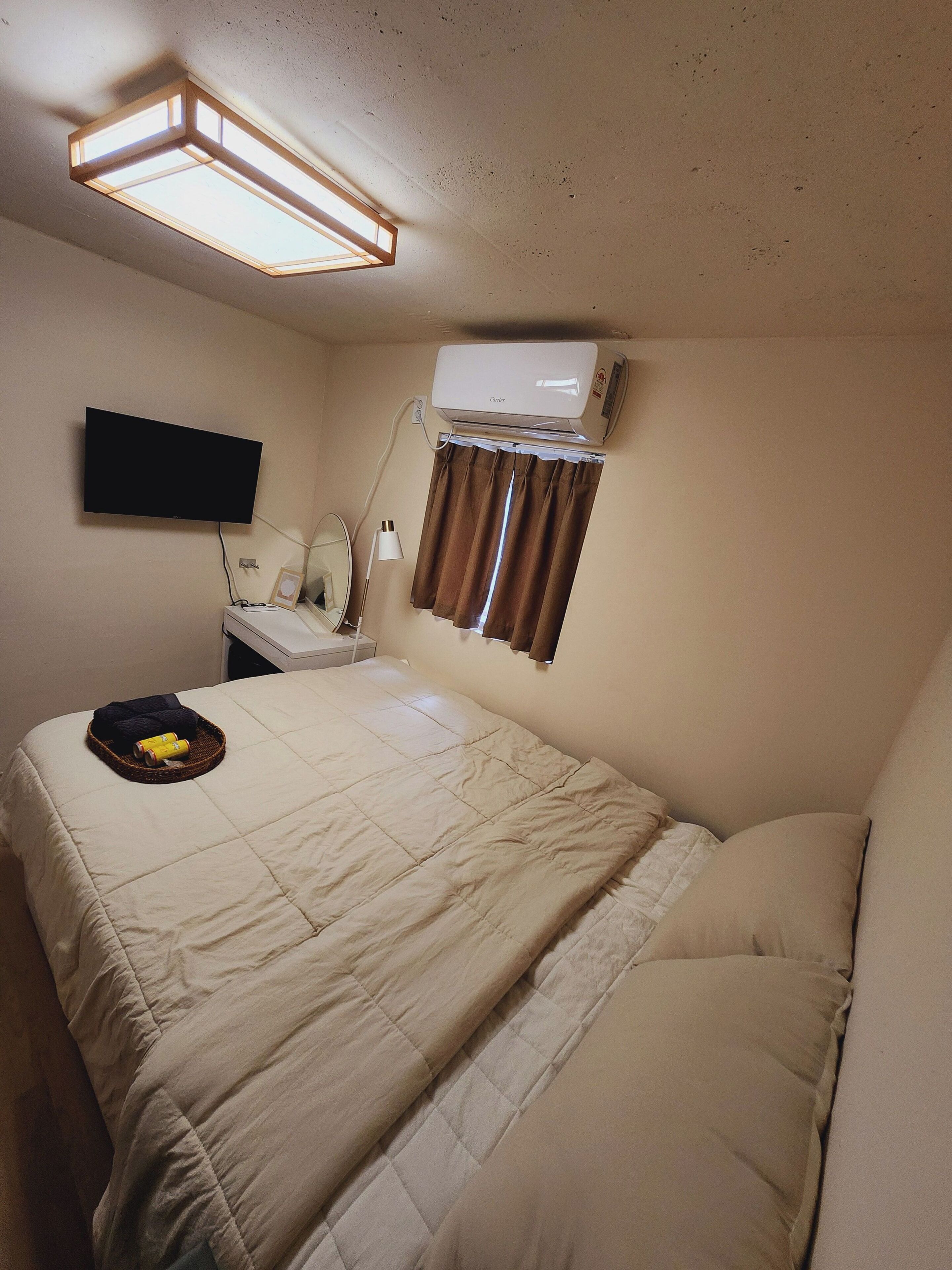 A hotel room with a bed, a flat-screen TV mounted on the wall, an air conditioner, a bedside table with a lamp, and a window with curtains.