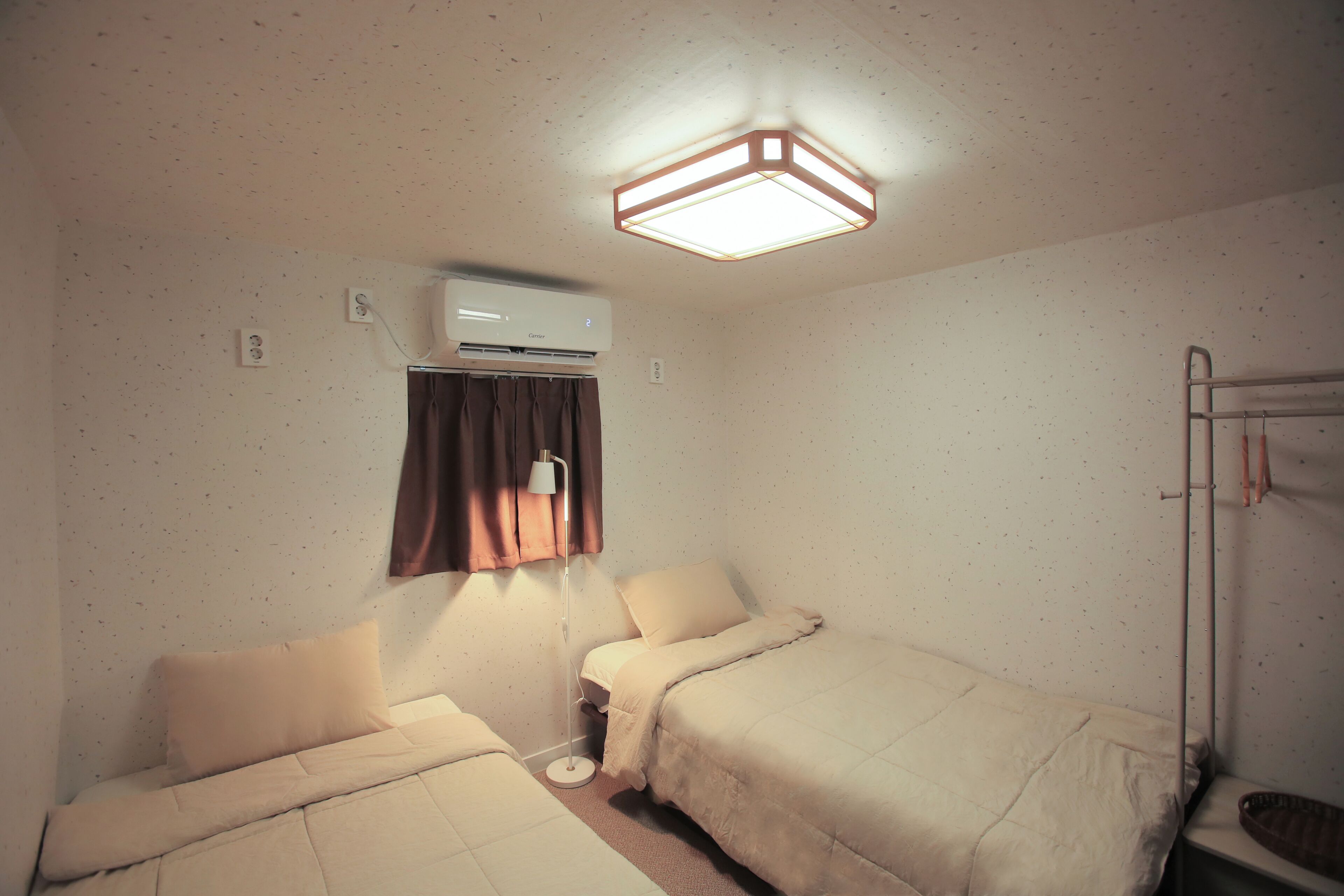 A small, simple room with two beds, a single bedside table, a lamp, an air conditioning unit, and a radiator.