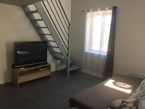 Ein Wohnzimmer mit Fernseher auf einem Holzboden, einer Treppe und einem Fenster mit transparenten Vorhängen.