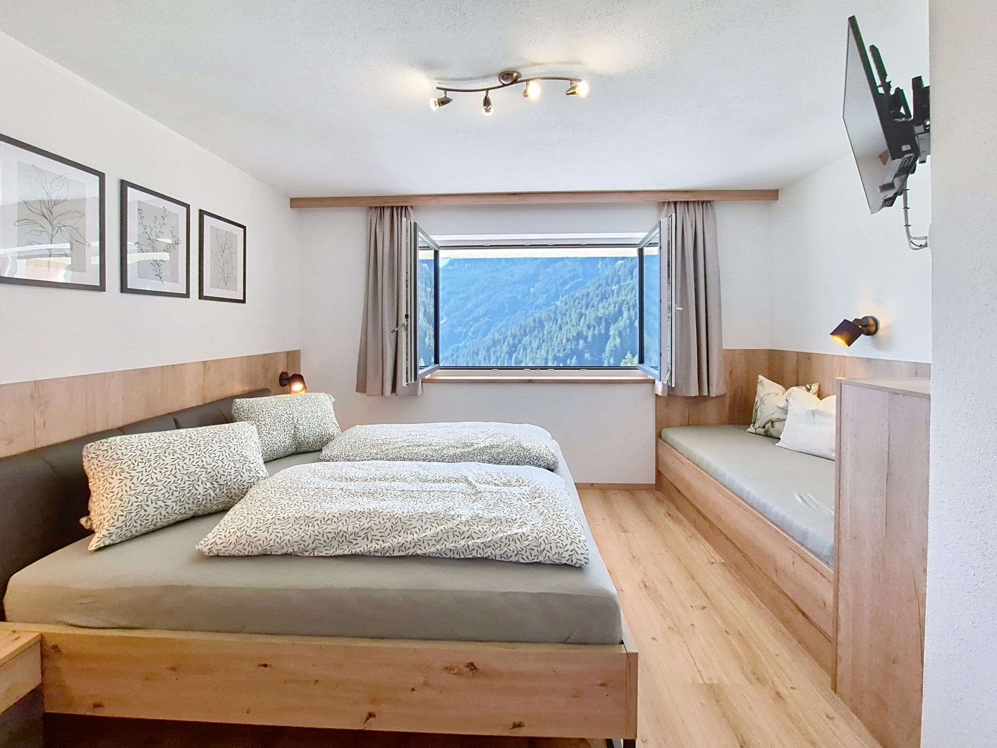 A bedroom with a bed, a window with a mountain view, a bedside table, and a wall-mounted TV.