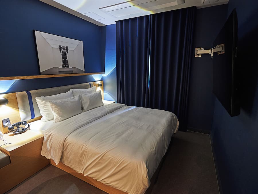 A hotel room with a bed, bedside table, lamp, artwork, curtains, and a TV.
