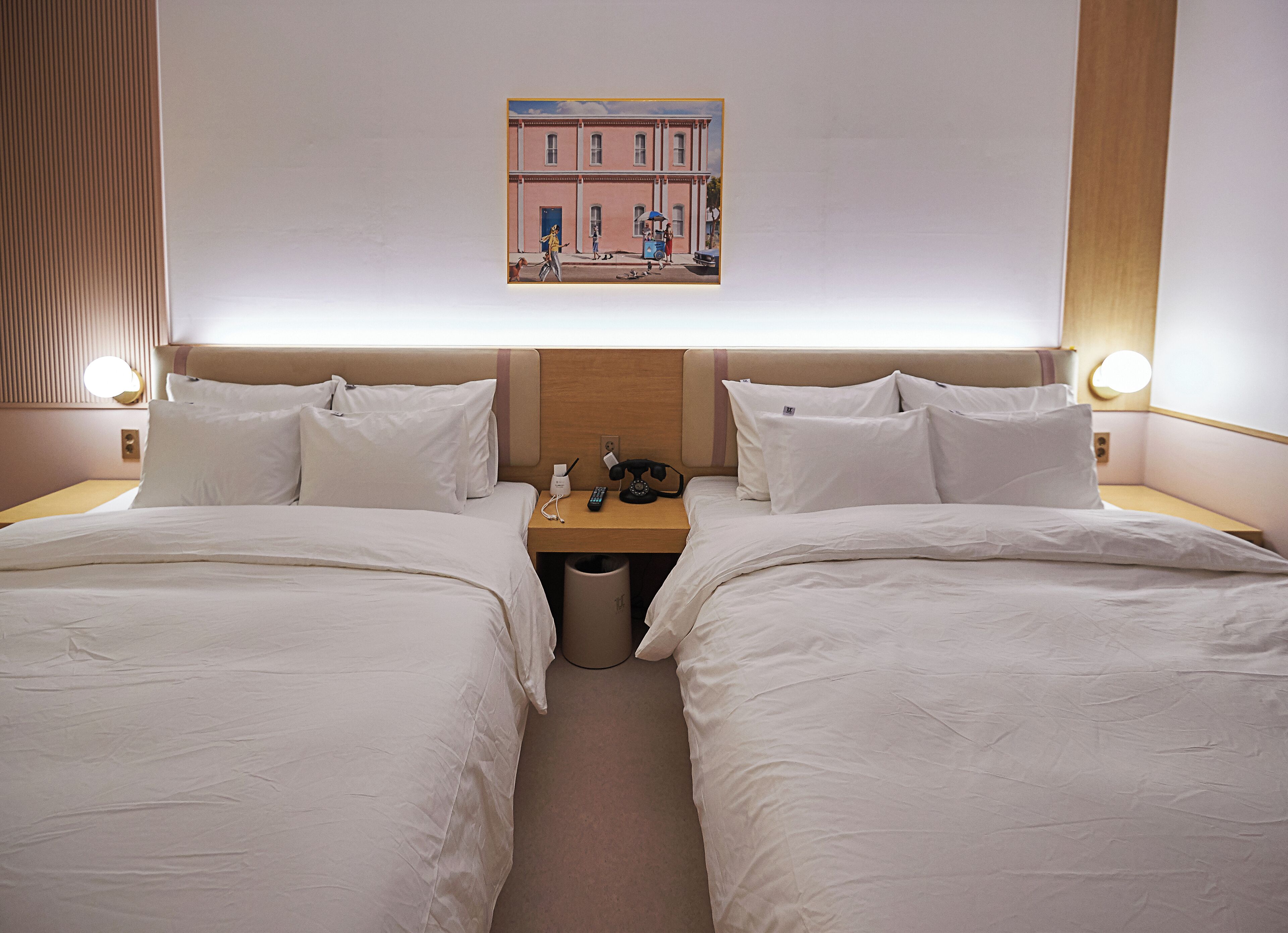 Habitación de hotel con dos camas, una mesita de noche con lámpara, un cesto de basura y un cuadro en la pared.