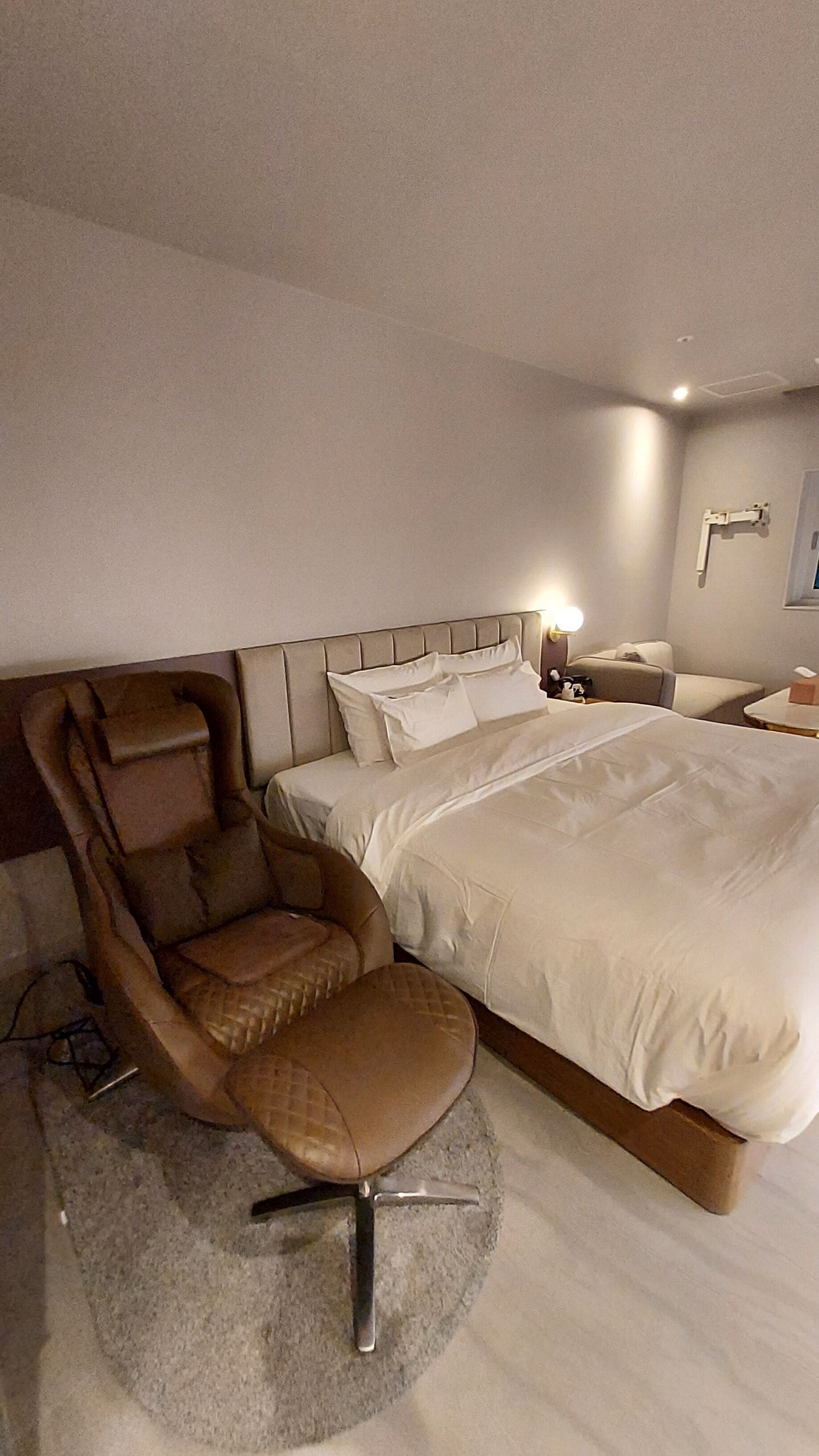 A modern hotel room with a large bed, a leather armchair, and a bedside table with a lamp.