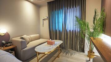 Habitación de hotel moderna con sofá, una mesita, lámpara de noche, planta en maceta y vista al exterior a través de una ventana con cortinas.