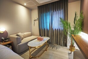 Habitación de hotel moderna con sofá, una mesita, lámpara de noche, planta en maceta y vista al exterior a través de una ventana con cortinas.