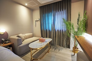 Quarto de hotel moderno com sofá, uma mesinha, abajur, planta em vaso e vista para o exterior através de uma janela com cortinas.