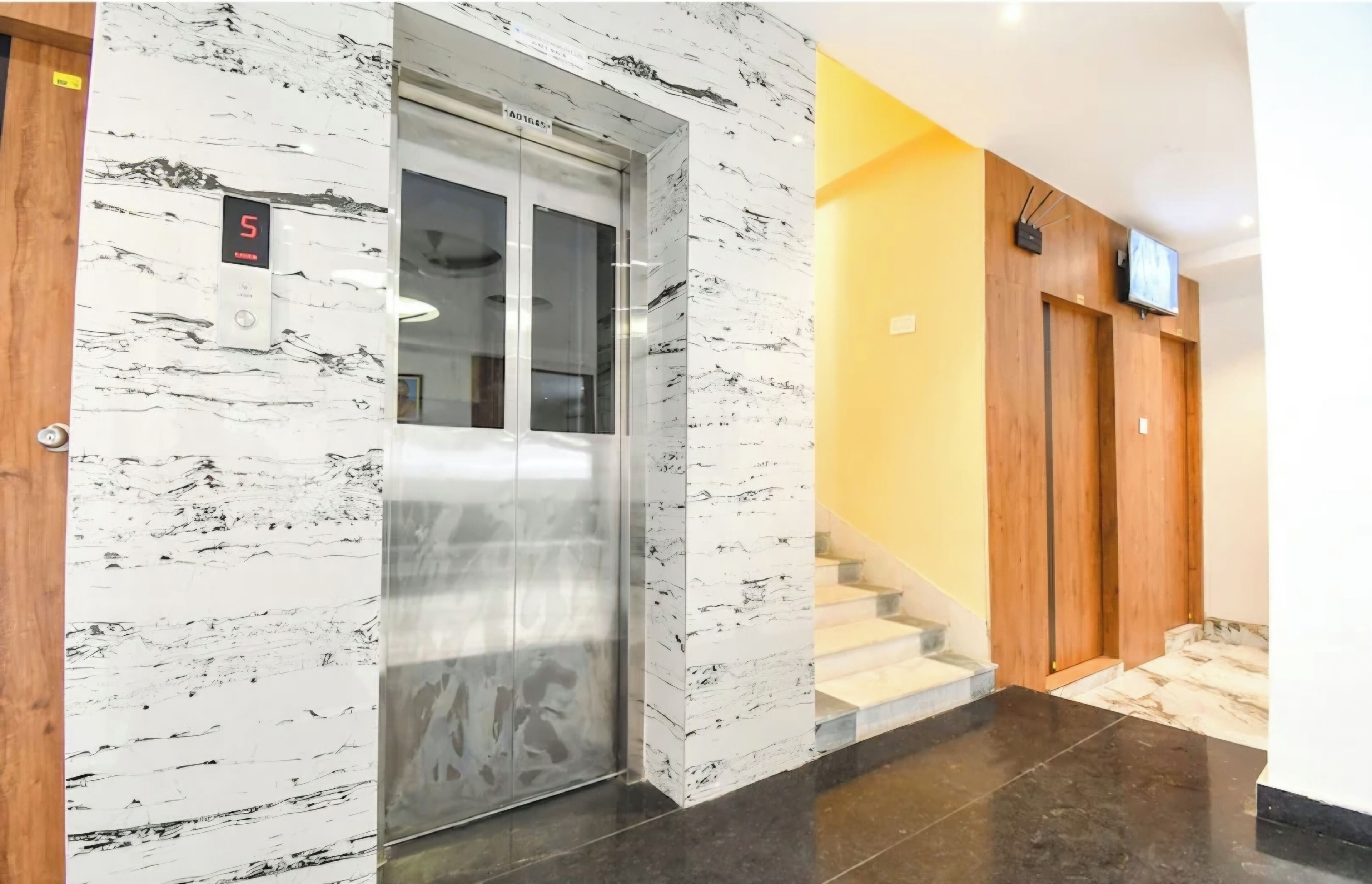An elevator with a digital display, marble walls, a wooden door, and a staircase with a yellow wall.