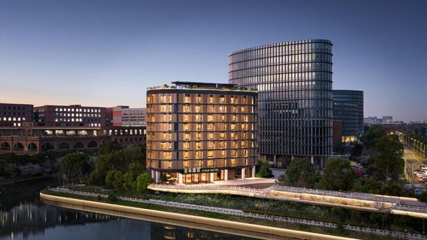 Un hotel moderno con un edificio circular y una estructura de cristal rectangular, ubicado junto a un río con un puente.