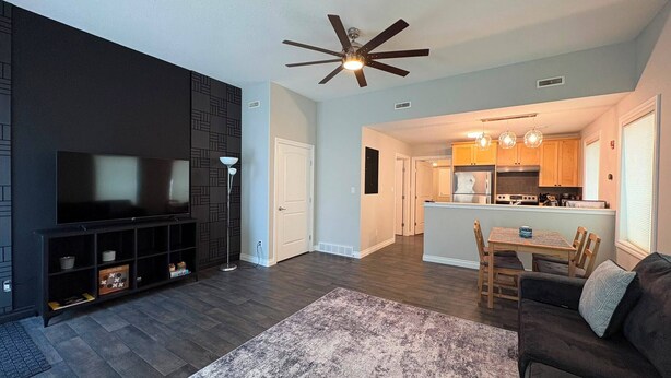 A modern living room with a flat-screen TV, dark wood flooring, a patterned rug, and a ceiling fan. - Central | World's Largest Dino & Splash Park 🌟 (Drumheller)