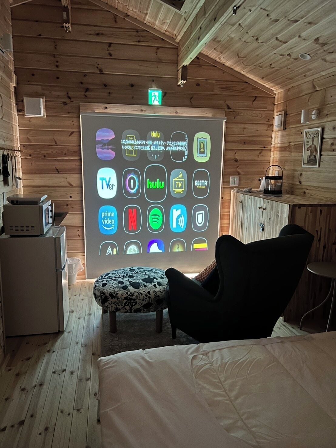 A cozy room with a wooden ceiling, a bed, a black chair, a small table, and a wall-mounted screen displaying various app icons.