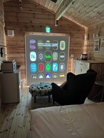 Habitación acogedora con techo de madera, una cama, una silla negra, una mesita y una pantalla en la pared que muestra varios iconos de aplicaciones.