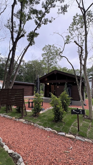 A rustic cabin with a porch, surrounded by trees and a gravel path. - mokku (Nasu)