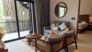 Ein modernes Hotelzimmer mit einer Couch, zwei Sesseln, einem runden Couchtisch, einem Bett, einem Spiegel und Blick durch eine Glastür.