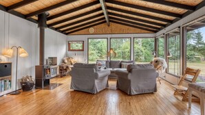Uma sala de estar ampla com pé direito alto, vigas de madeira, lareira e grandes janelas com vista para o exterior.