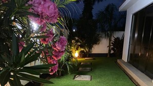 Un césped bien cuidado con pasto artificial, un camino y un pequeño jardín con flores y plantas.
