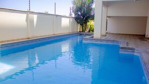 Una piscina con bordeado de baldosas, rodeada por un muro blanco y un árbol con flores rosadas.