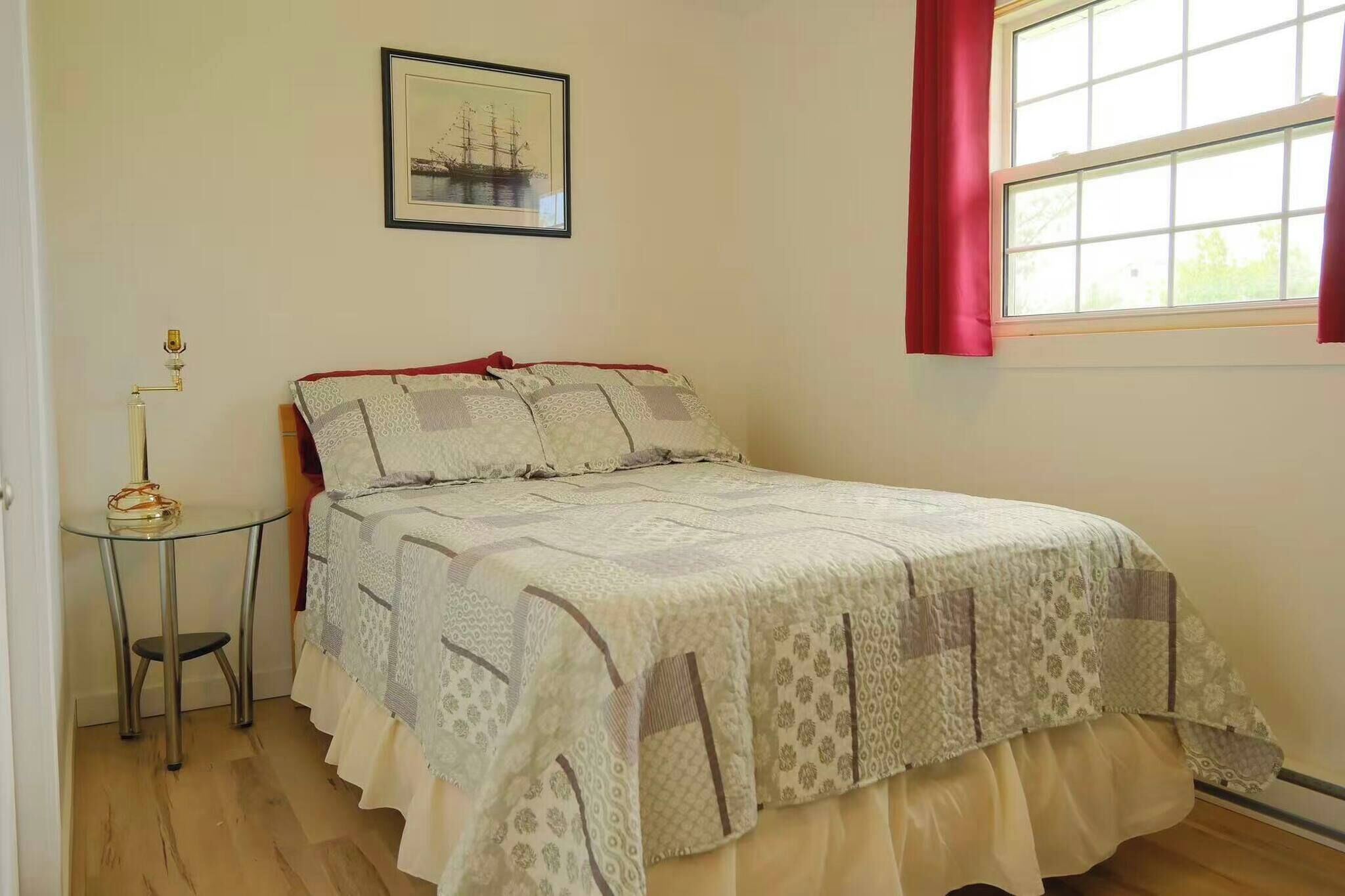 Un dormitorio con cama, una mesita, una lámpara, una ventana con cortinas y un cuadro en la pared.