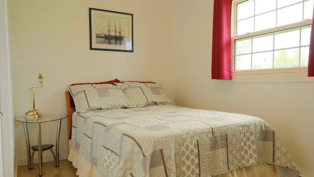 A bedroom with a bed, a small table, a lamp, a window with curtains, and a framed picture on the wall.