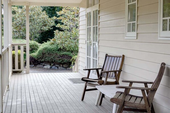 A wooden deck with two Adirondack chairs and a small table, a white railing, and a view of a garden with various plants and a stone wall. - Mill Hill Cottage - Montville (Sunshine Coast)