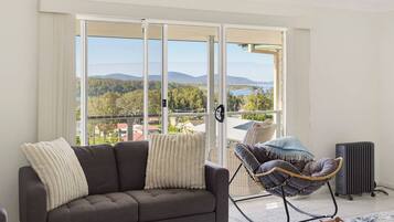 A modern living room with a grey sofa, a wicker chair, and a view of a lake and mountains through large glass doors.