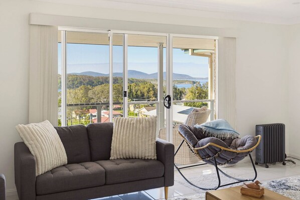 Un salon moderne avec un canapé gris, un fauteuil en osier et une vue sur un lac et des montagnes, visible à travers de grandes portes vitrées.