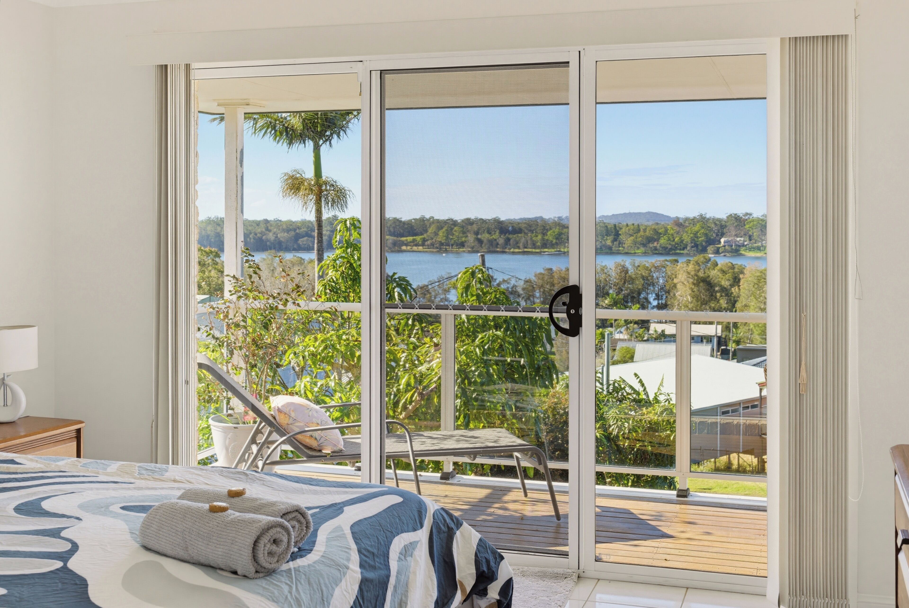 A bedroom with a large sliding glass door leading to a balcony with a view of a lake and trees.