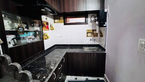 A compact kitchen with dark wood cabinets, a black countertop, white subway tiles, and a small window. - The Trikut Homestay – Just 1.4 km from Gadisar Lake (Jaisalmer)