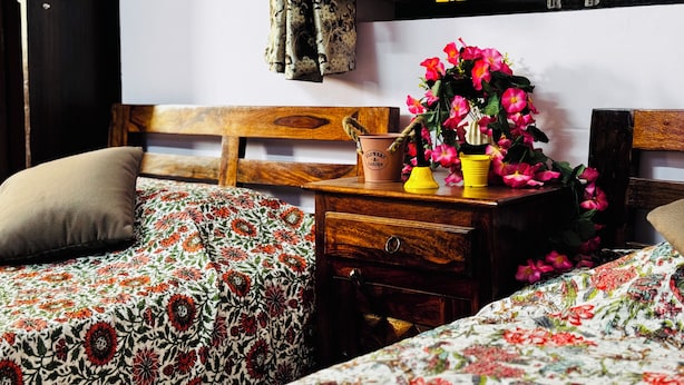A bedroom with a wooden headboard, floral bedding, a nightstand with a potted plant, and a window with yellow blinds. - The Trikut Homestay – Just 1.4 km from Gadisar Lake (Jaisalmer)