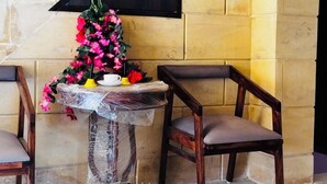 A room with a stone wall, a small table with a plastic cover, two wooden chairs, and a decorative arrangement of flowers and a teapot. - The Trikut Homestay – Just 1.4 km from Gadisar Lake (Jaisalmer)