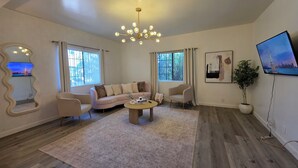 Ein Wohnzimmer mit einer Couch, Sesseln, einem Couchtisch, einer Deckenleuchte, einem großen Spiegel und einem Fernseher.