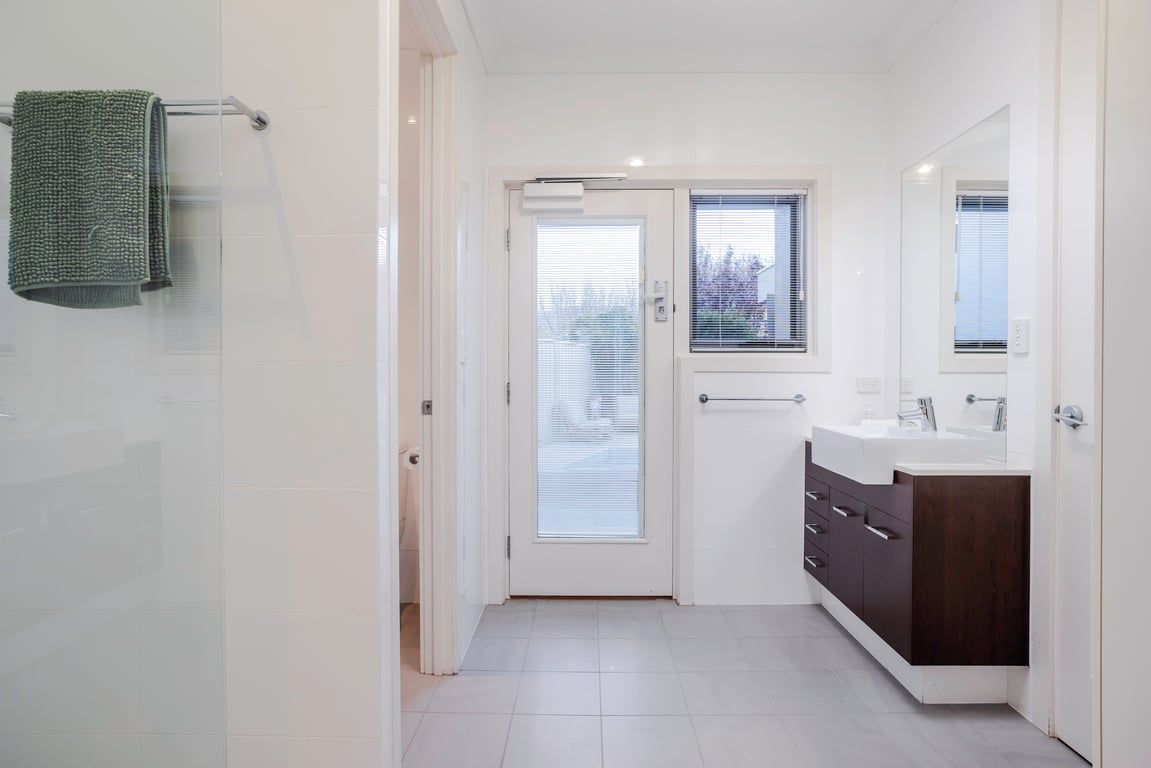 A modern bathroom with a glass shower enclosure, a white door, a window with blinds, and a dark wood vanity with a mirror.
