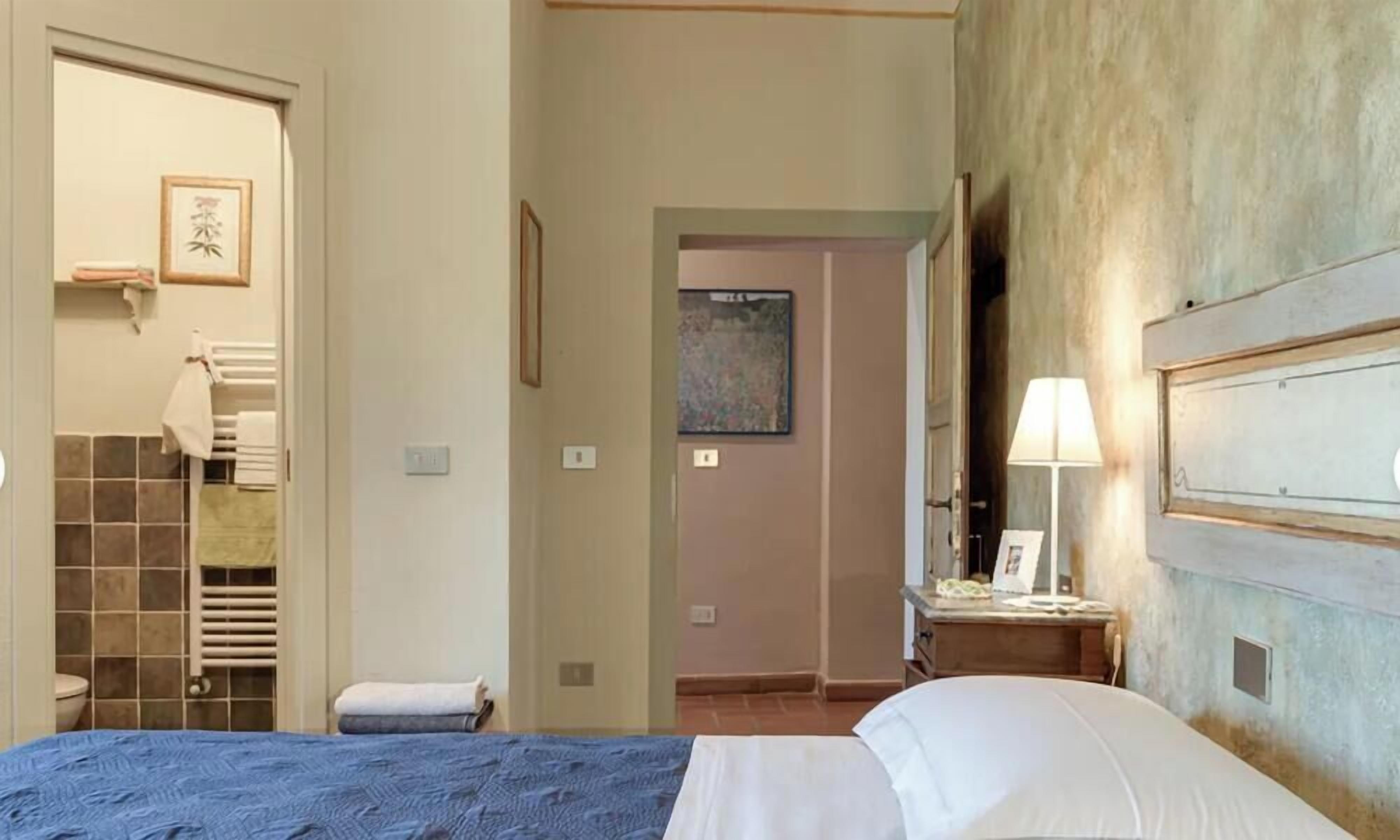 A bedroom with a bed, a bedside table with a lamp, a bathroom with a towel rack, and a door leading to another room.