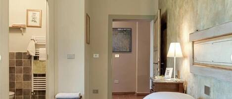 A bedroom with a bed, a bedside table with a lamp, a bathroom with a towel rack, and a door leading to another room.