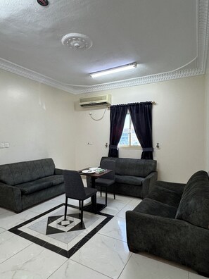 A living room with a sofa set, a dining table with chairs, a patterned rug, and an air conditioning unit on the wall.