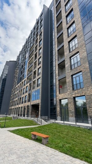 A multi-story residential building with a modern architectural design, featuring balconies, large windows, and a paved walkway with a bench.