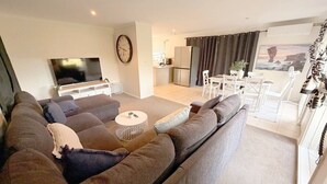 A modern living room with a large sofa, a flat-screen TV, a dining area with a table and chairs, and a kitchen in the background.