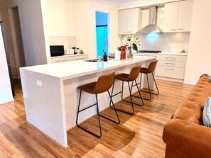 A modern kitchen with a central island, bar stools, and a wooden floor. - Luxury 5BR+5Bath Home •Style, Space & Comfort • EV (Truganina)