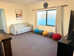 A living room with a sofa, a window with curtains, a clock, and colorful knitted poufs. - Luxury 5BR+5Bath Home •Style, Space & Comfort • EV (Truganina)