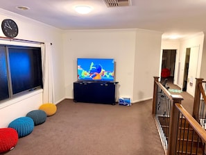 A living room with a flat-screen TV on a stand, a window with curtains, a staircase with a railing, and a carpeted floor. - Luxury 5BR+5Bath Home •Style, Space & Comfort • EV (Truganina)