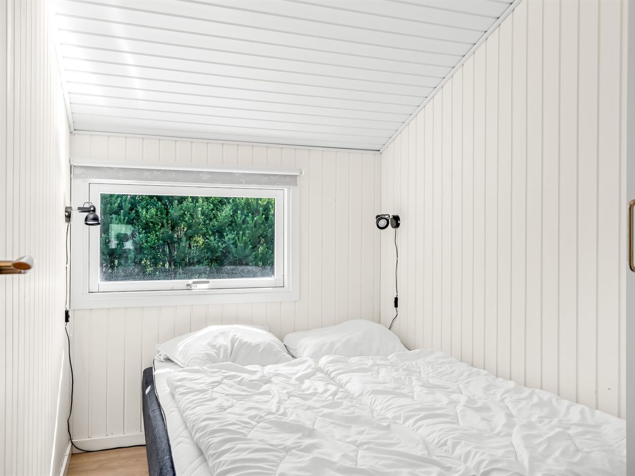 Een slaapkamer met een bed, een raam met uitzicht op groen en een wandlamp.