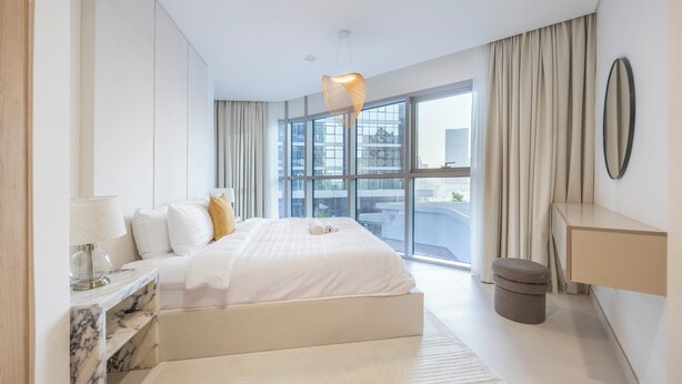 Ein modernes Hotelzimmer mit einem großen Bett, einem мраморным Nachttisch, einem kleinen Hocker, einem großen Fenster mit Vorhängen und einer hängenden Leuchte.