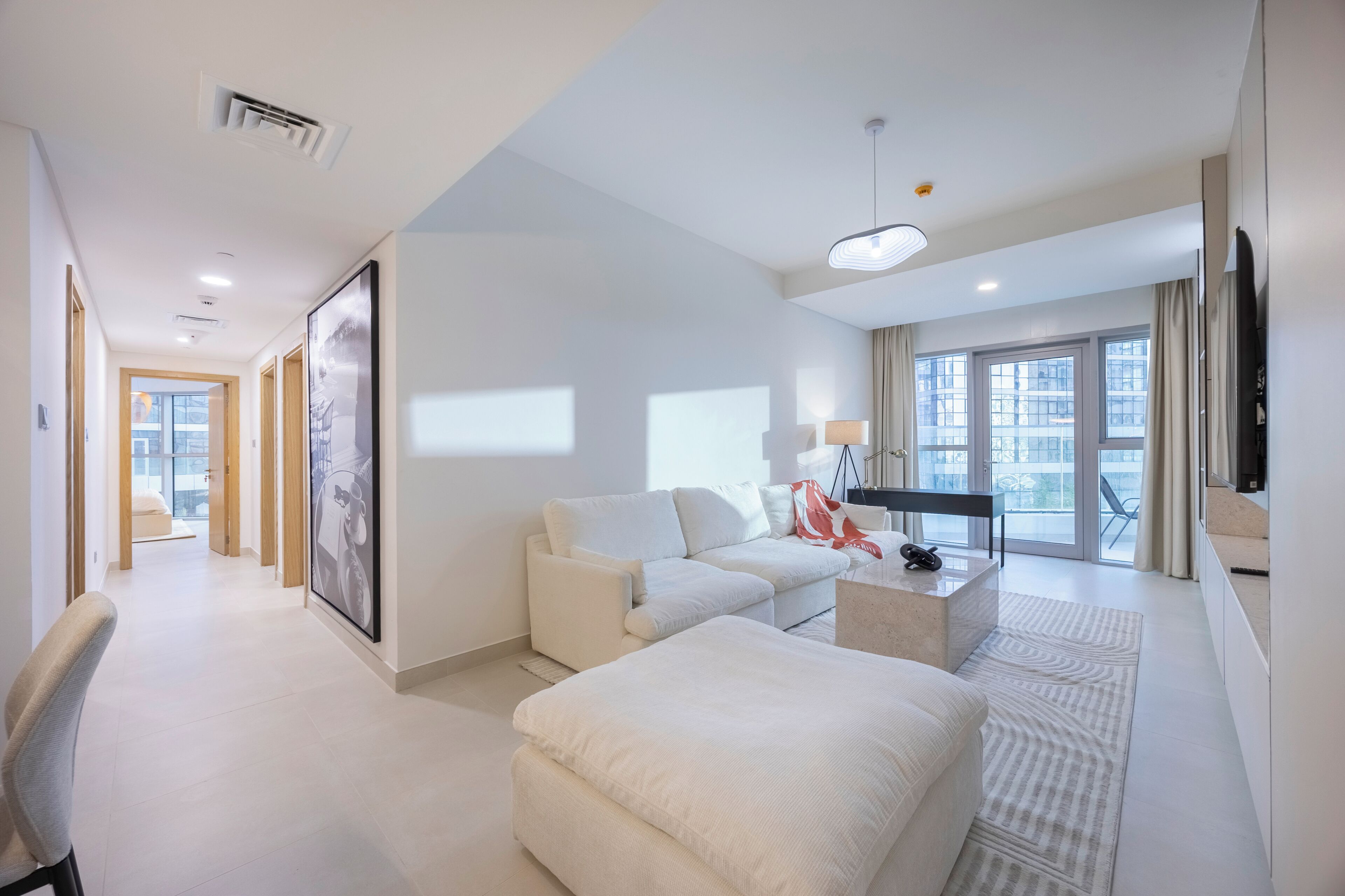 A modern hotel room with a sofa, ottoman, a desk with a lamp, a flat-screen TV, and large windows with curtains.
