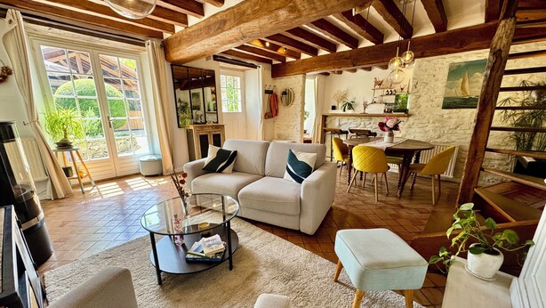 A spacious living area with a sofa, coffee table, and ottoman, wooden beams on the ceiling, and a dining area with chairs and a table. - La Maison des Roses - A charming house with a garden (Rully)