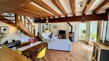 A rustic interior with exposed wooden beams, a dining area with a piano, a living room with a sofa, and a stone wall.